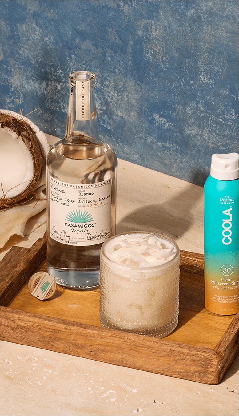 Casamigos Blanco Tequila bottle with a creamy coconut cocktail and COOLA SPF 30 sunscreen spray, styled with coconut and wooden accents on a sandy beige and blue background.