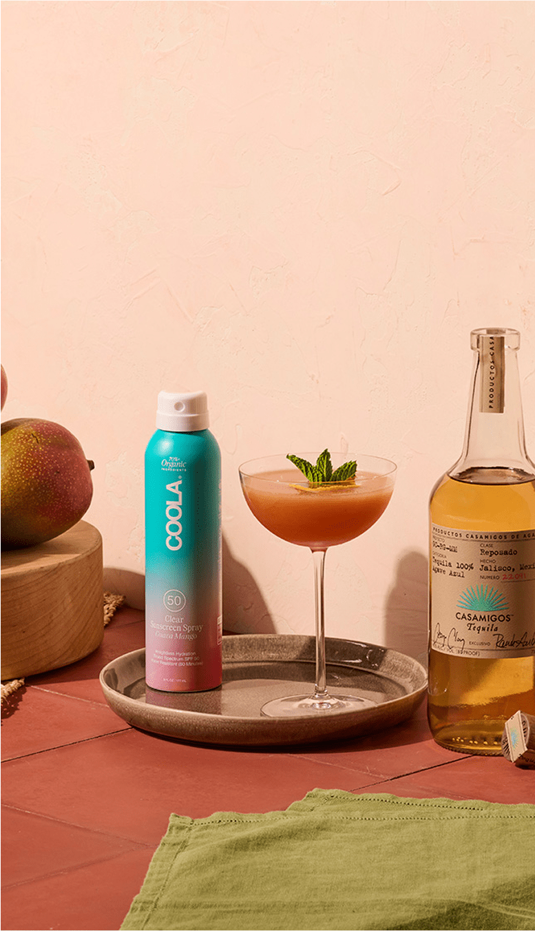 Casamigos Reposado Tequila bottle with a vibrant pink cocktail garnished with mint, accompanied by COOLA SPF 30 sunscreen spray, mango, and a neutral pink backdrop.