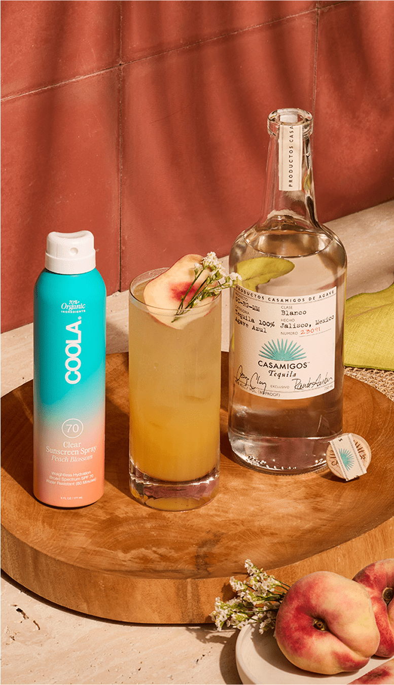 Casamigos Blanco Tequila bottle with a fresh cocktail garnished with herbs and peach slices, paired with COOLA SPF 70 sunscreen spray on a warm terracotta background.