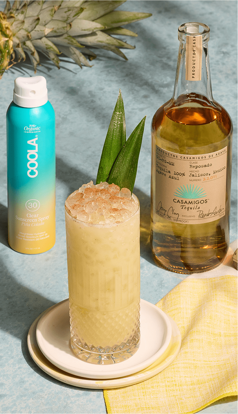 Casamigos Reposado Tequila bottle with a tropical cocktail garnished with pineapple, alongside COOLA SPF 30 sunscreen spray on a pastel blue background.