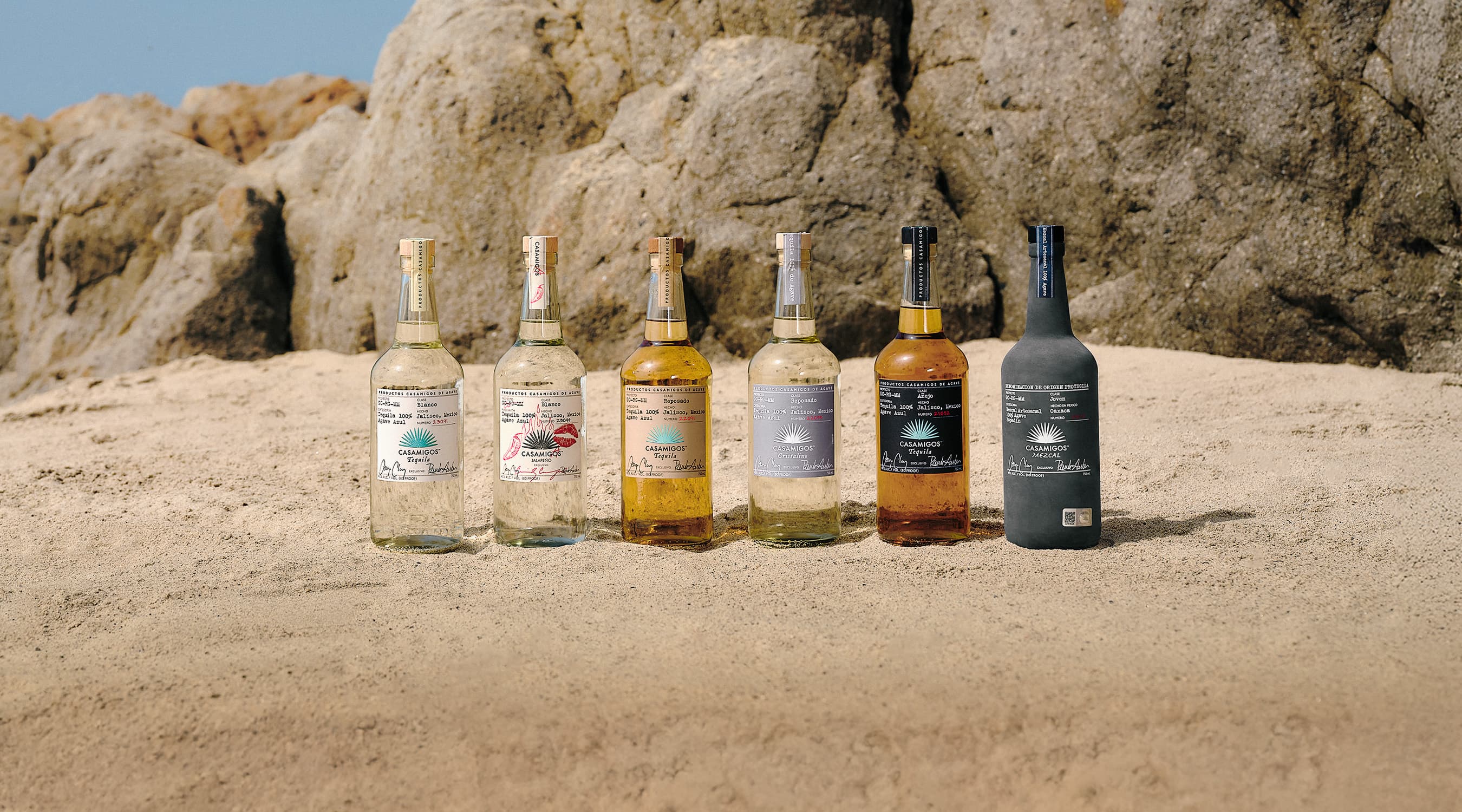 Six Casamigos bottles, including Blanco, Reposado, Añejo tequilas, and Casamigos Mezcal, displayed upright on a sunlit sandy beach with large rocks in the background.